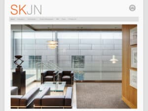SKJN Architekten website. A screen capture of a website page with a clean, white, minimalist design in white and light grey with a simple, straight character font for the title "SKJN", of which the first two letters are orange and the second two grey, below that a navigation bar with some links in grey and across the rest of the page is the masthead photo of a clean, very modern building interior with square-shaped chairs and a wood finish was visible near a metal staircase